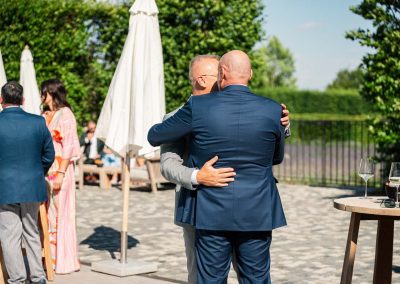 Esküvői kép két férfi öleléssel, nyári kerthelyiségben.