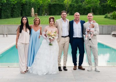 Esküvői csoportkép a medencénél, boldog pár és családtagok.