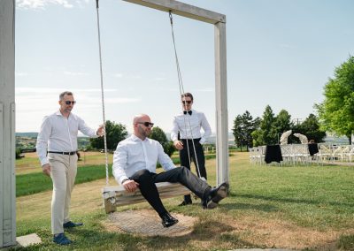 Esküvői férfiak a szabadban, egy hintán, elegáns öltözetben, napos időben.