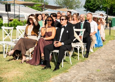 Esküvői vendégek ülnek a szabadban, elegáns ruhában, napfényes időben.