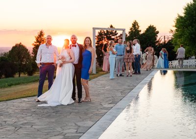 Esküvői vendégek a naplementében a szabadban, elegáns ruhában.