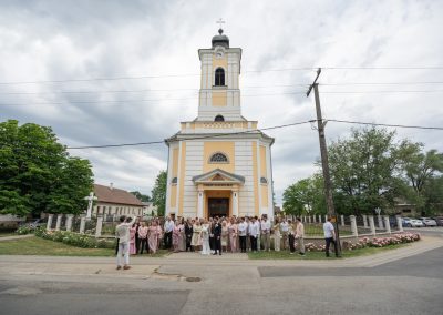 Esküvői csoportkép a templom előtt, boldog pár és vendégek a szertartás után.