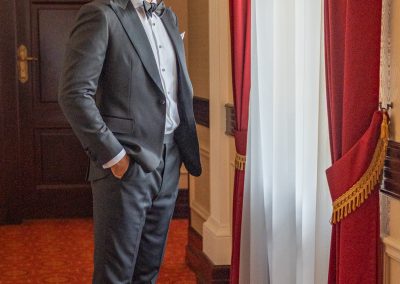 Elegáns férfi esküvői öltönyben, szobában, vörös szőnyeggel és függönyökkel.